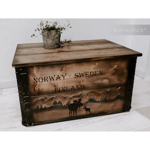 May include: A wooden storage chest with black metal accents. The chest is decorated with a mountain scene and two moose silhouettes. The text "NORWAY - SWEDEN FINLAND" is printed on the front of the chest.