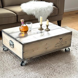 Peut inclure: Une table basse en bois rustique avec une finition vieillie, des ferrures métalliques et des roulettes. La table contient une bouteille de whisky, un verre et deux bougeoirs avec des bougies allumées. Le texte "Old Scotch Whisky Rare Mill" est visible.