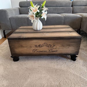 Könnte beinhalten: Quadratischer Couchtisch aus dunkel gebeiztem Holz auf schwarzen Rollen. Der Tisch trägt den Schriftzug "Premiere Cuvée" und ein dekoratives Design. Eine weiße Vase mit weißen und rosa Blumen steht darauf.