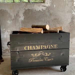 Op de afbeelding: Een zwarte houten kist op wielen, gevuld met brandhout. De kist heeft gouden letters met de tekst "CHAMPAGNE Premiere Cuvée". De kist heeft een rustieke uitstraling.