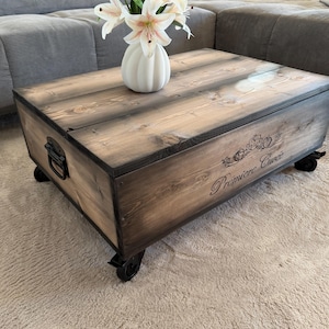 Könnte beinhalten: Ein rustikaler Couchtisch aus Holz mit dunkler Beize, mit einem Klappdeckel und Metallbeschlägen. Der Tisch steht auf kleinen Rollen und trägt den Aufdruck "Premiere Cure". Eine weiße Vase mit weißen und rosa Blumen steht darauf.