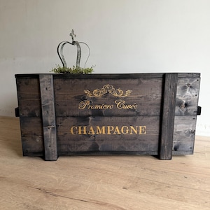 Könnte beinhalten: Eine dunkel gebeizte Holzkiste für Champagner mit goldfarbenen Buchstaben, die "Premiere Cuvée CHAMPAGNE" lauten. Die Kiste hat schwarze Metallgriffe und ist mit einer Metallkrone und Grün dekoriert.