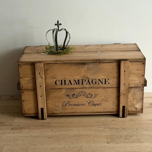 May include: A wooden champagne crate with the word "CHAMPAGNE" printed on the front. The crate is light brown and has a decorative metal crown on top. The crate is made of wood and has a rustic appearance.