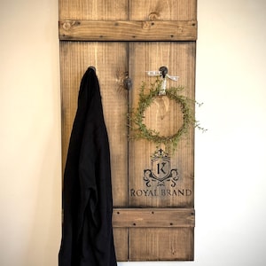 Peut inclure: Un porte-manteau mural en bois rustique avec une teinture foncée. Il comporte deux crochets en métal, une petite couronne verte et le texte "ROYAL BRAND" avec un logo de crête. Un vêtement noir est suspendu à un crochet.