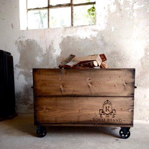 Könnte beinhalten: Eine rustikale Holz-Aufbewahrungsbox auf Rädern, gefüllt mit Brennholz. Die Box ist in einem warmen Braunton gehalten und trägt das Logo "Royal Brand". Die Box befindet sich in einem Raum mit einem Fenster.