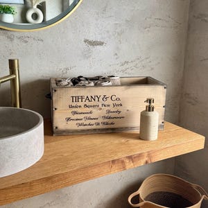 Op de afbeelding: Een houten kist met de tekst "TIFFANY & Co." en andere tekst, samen met een zeepdispenser, staat op een houten plank. Een geweven mand staat eronder. De kist bevat opgerolde handdoeken. De achtergrond is een lichtgrijze muur.