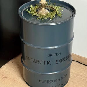 Può includere: Un barile di metallo blu scuro con la scritta "BRITISH ANTARCTIC EXPEDITION BURROUGH & CO LONDON". Il barile ha un coperchio con un piccolo portacandele e una ghirlanda di finta vegetazione. La candela è beige.