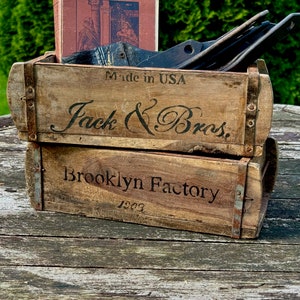Può includere: Due casse di legno impilate e usurate con fasce metalliche. La cassa superiore recita "Jack & Bros." e "Made in USA". La cassa inferiore dice "Brooklyn Factory 1903". Un libro vintage e oggetti neri sono all'interno.