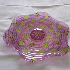 May include: A pink glass serving dish with a scalloped edge and a concentric circle design. The dish is decorated with green and gold dots.