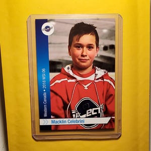 Może przedstawiać: Karta sportowa ze zdjęciem młodego hokeisty w czerwonej koszulce z białymi akcentami. Karta zawiera napis Selects, Western Canada 2018 WSI 06 oraz imię i nazwisko Macklin Celebrini wydrukowane w kolorze niebieskim i białym.