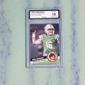 May include: A graded Arch Manning 2020 Hot Shot Prospects trading card. The card features a color image of a football player in a green and white uniform throwing a football. The card is encased in a clear plastic holder.
