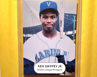 May include: A baseball card of Ken Griffey Jr. in a Mariners uniform, holding a bat. He wears a blue cap with a white "V" and a white and blue jersey. The card is in a protective case, with text that reads "KEN GRIFFEY JR. Eastern League Prospects."