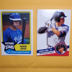 Pode incluir: Dois cartões de beisebol colecionáveis ​​com Aaron Judge. O primeiro cartão é da série 2011 Alaska League Prospects e mostra Judge com um uniforme azul e um taco. O segundo cartão é da série 2014 Fall League Prospects e mostra Judge com um uniforme do New York Yankees rebatendo. Ambos os cartões têm o texto "FUTURE STARS" e "AARON JUDGE".