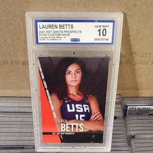 May include: A graded trading card featuring Lauren Betts, a basketball player, in a USA jersey. The card is encased in a protective holder and has a GEM MINT 10 grade. Text on the card includes "2021 HOT SHOTS PROSPECTS" and "ACEO CUSTOM ISSUE."