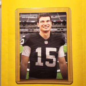 Puede incluir: Una tarjeta coleccionable de Fernando Mendoza, novato del draft de 2026. La tarjeta muestra a Mendoza con un uniforme de fútbol en blanco y negro con el número 15. La tarjeta está en una funda protectora sobre un fondo amarillo.
