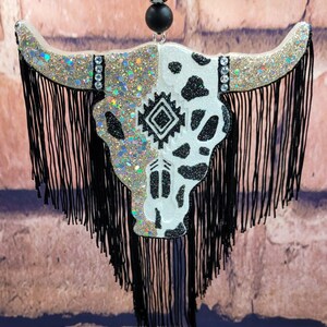 May include: A black and white cow skull with a geometric pattern and glitter accents. The skull is decorated with black fringe and a black and white beaded necklace.