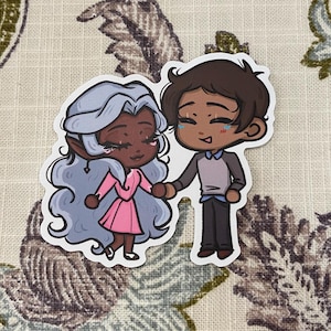 May include: A die-cut sticker featuring two cartoon characters holding hands. One character has gray hair and wears a pink dress, while the other has brown hair and wears a gray shirt and black pants. The sticker has a white border and is on a floral background.