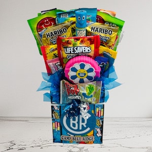 May include: A candy bouquet featuring a variety of colorful candies and treats. The arrangement includes Haribo Goldbears, Life Savers Gummies, and Ring Pop Mysteries. The base is a box with "CAMP BLUE RIDGE" text and a blue and white logo.