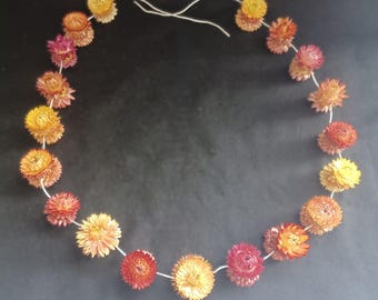 May include: A circular garland or necklace constructed from a thin, flexible silver-toned wire strung with numerous dried strawflowers in various shades of yellow, orange, red, and pink, arranged in a repeating pattern along the wire.