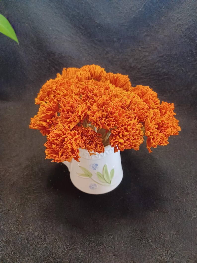 May include: A bouquet of dried orange marigold flowers in a white ceramic pitcher with a blue floral design.