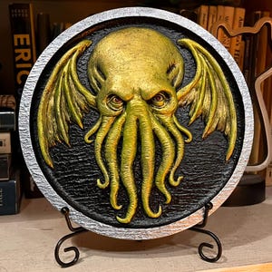 May include: A round, textured wall plaque featuring a golden-yellow Cthulhu design with wings and tentacles, set against a black background. The plaque has a silver border and is displayed on a black metal stand, showcasing a unique, fantasy-themed art piece.
