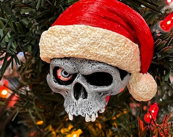 May include: A white skull wearing a red Santa hat with white fur trim. The skull has one visible eye with a red iris. The ornament is hanging from a Christmas tree.