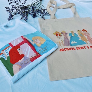 Peut inclure: Un t-shirt blanc et un sac fourre-tout beige avec des illustrations colorées. Le t-shirt présente un graphisme de deux femmes avec du texte en français. Le sac fourre-tout affiche un groupe de femmes et le texte "JACQUES DEMY'S GANG" en rouge.