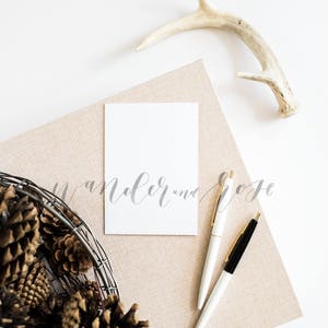 May include: A white card with the text "Wander and Rose" written in cursive on a beige textured surface. Two pens, one black and one white, are on the surface. A basket of pine cones is in the lower left corner of the image. A white antler is in the upper right corner of the image.