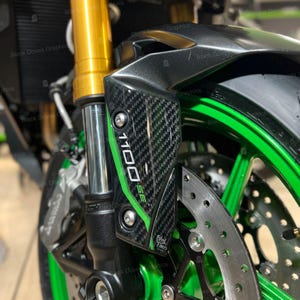 Può includere: Primo piano della parte anteriore di una moto. L'immagine presenta un elemento decorativo nero e in fibra di carbonio con la scritta "1100 SE" in bianco e verde. La moto ha una ruota verde e componenti delle sospensioni dorati.