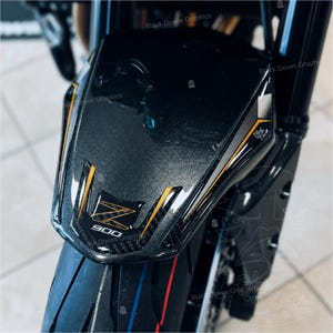 Pu&ograve; includere: Primo piano di un parafango anteriore di una moto. Il parafango &egrave; grigio scuro con accenti dorati e neri. Il testo "900" e una "Z" stilizzata sono visibili. Lo pneumatico ha strisce rosse e blu.