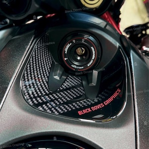Può includere: Primo piano dell'area del serbatoio del carburante di una moto. Il design presenta un motivo in fibra di carbonio e un interruttore di accensione nero con dettagli rossi. Il testo "BLACK DOVES GRAPHICS" è visualizzato in rosso.
