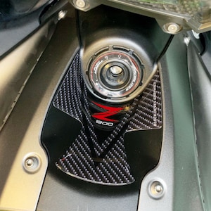 Può includere: Primo piano di un serbatoio di benzina per moto nero con un motivo in fibra di carbonio e un logo "900" nero e rosso. Il serbatoio ha un tappo nero con un'etichetta argentata "IMMOBILIZER".