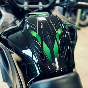 Può includere: Serbatoio nero per moto con un design geometrico verde e oro. Il design include il numero "1100" e il testo "Black Doves Graphics". Il serbatoio ha una finitura lucida.
