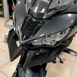 Op de afbeelding: Close-up van de voorkant van een donkergrijze motorfiets. De motorfiets heeft een strak ontwerp met een zwarte grille, heldere koplampen en een getinte voorruit. Het Kawasaki-logo is zichtbaar.