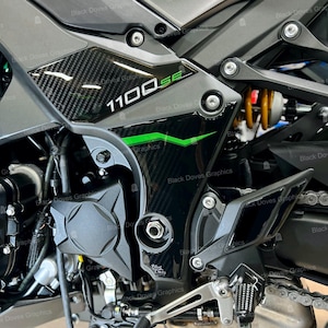 Può includere: Primo piano della carrozzeria nera e grigia di una moto, con dettagli in fibra di carbonio e la scritta "1100SE" in bianco e verde. Il design include una striscia verde e vari componenti meccanici.