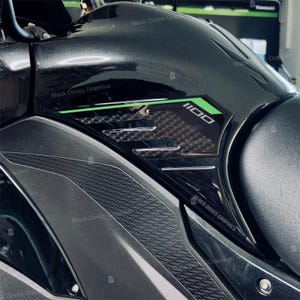 Può includere: Primo piano della carrozzeria nera e grigia di una moto. Il design include un motivo strutturato e il numero "1100" in bianco con un bordo verde. Il testo "Black Doves Graphics" è visibile.