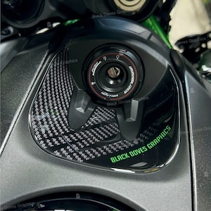 Può includere: Primo piano di un serbatoio di benzina per moto nero con un motivo in fibra di carbonio e un adesivo verde "BLACK DOVES GRAPHICS". Il serbatoio ha un coperchio nero per la chiave di accensione con le parole "PUSH" e "IMMOBILIZER" stampate su di esso.