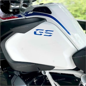 以下が含まれることがあります： 青と白のアクセントが付いた白いオートバイの燃料タンク。「GS」の青い文字が側面にあります。「Black Doves Graphics」の文字がタンクに繰り返されています。