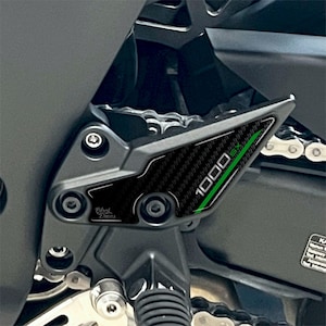 以下が含まれることがあります： 黒いオートバイのフットペグプロテクター。カーボンファイバーパターンと緑色の「1000 SX」の文字が特徴です。プロテクターには「Black Daces」の文字も表示されています。