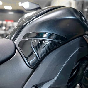 Può includere: Primo piano del serbatoio nero di una moto con un inserto in fibra di carbonio. Il numero "1100" è visualizzato in bianco. Il serbatoio ha una finitura lucida ed è parzialmente visibile.