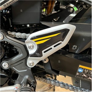 Peut inclure: Gros plan sur un protège-chaîne de moto avec un design noir et blanc. Le protège-chaîne a une bande jaune et le texte "Black Doves" imprimé dessus.