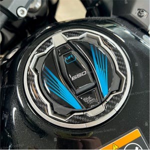 Pode incluir: Uma tampa de tanque de gasolina de motocicleta preta com um design de fibra de carbono e um acento azul e prateado. A tampa tem um botão de elevação e o texto "650" e "Black Doves" impresso nela.