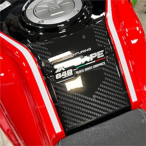 Puede incluir: Un tanque de gasolina de motocicleta rojo con un panel de fibra de carbono negro y una calcomanía plateada que dice "XAPE ADVENTURING 649 BLACK DOVES GRAPHICS".