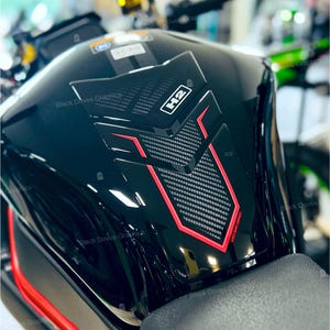 Può includere: Un serbatoio del carburante nero lucido per moto con un pannello centrale strutturato in stile fibra di carbonio. Accenti rossi delineano il pannello e altri elementi di design. Il testo "H2" è visibile sul pannello. Le parole "Black Doves Graphics" sono anch'esse visibili.