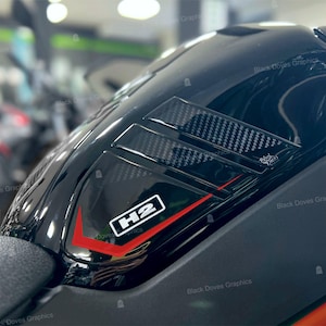Può includere: Primo piano di un serbatoio di moto nero lucido. Include prese d'aria in stile fibra di carbonio e una grafica bianca e rossa con la scritta "H2". Il serbatoio ha un design elegante e aerodinamico.