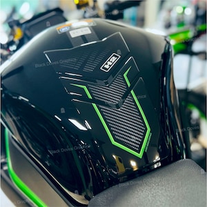 Può includere: Serbatoio del carburante per moto nero con un design in fibra di carbonio verde e nero. Il serbatoio ha un adesivo che dice "H2" e "Black Doves Graphics".