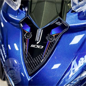 May include: Close-up of a blue motorcycle fairing with a black and carbon fiber accent piece. The accent piece has the number "1100" in white and is outlined with orange and blue accents. The text "Black Doves Graphics" is visible.