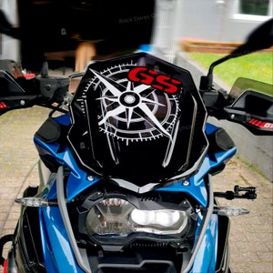 Könnte beinhalten: Ein blaues Motorrad mit einer schwarzen Windschutzscheibe, die mit einem Kompassdesign und den Buchstaben "GS" in Rot verziert ist. Das Motorrad hat einen schwarzen Scheinwerfer und einen schwarzen Lenker.