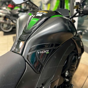 Peut inclure: Gros plan sur un réservoir de moto gris mat. Il présente des détails noirs et verts, dont le texte "1100 SE". Le design intègre des éléments en fibre de carbone et la lettre "Z".