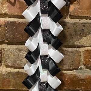 Op de afbeelding: Een decoratieve afstudeermum met afwisselende zwarte en witte satijnen linten. De mum heeft glitteraccenten en wordt gepresenteerd tegen een bakstenen muur. Een feestelijk accessoire.